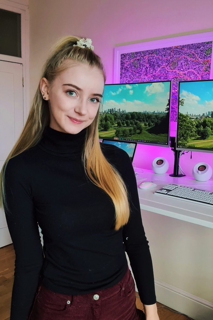 Holly standing in front of her computers with pink lights. She is wearing a black turtle neck and her hair is in a scrunchie