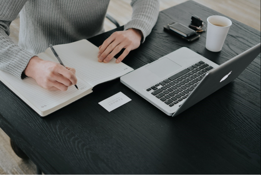 Person writing in a notebook with a laptop and coffee on a table