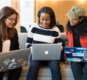 Three women sat on a couch looking at their laptops with coding stickers on them