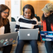 Three women sat on a couch looking at their laptops with coding stickers on them