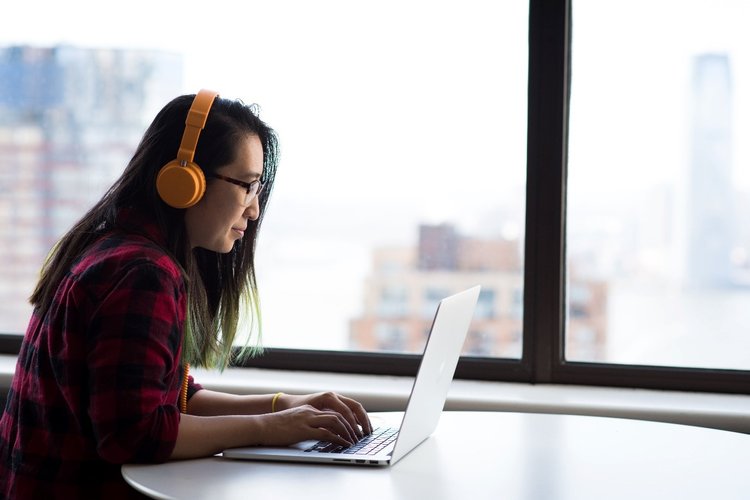 Woman sat at a desk listening to headphones working on a laptop in front of a window