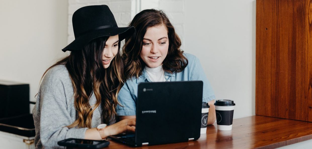 Two women looking at a laptop on a table top bench with coffee