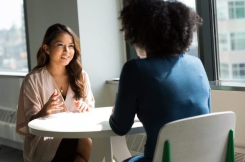 Two women speaking to each other over a table by a window