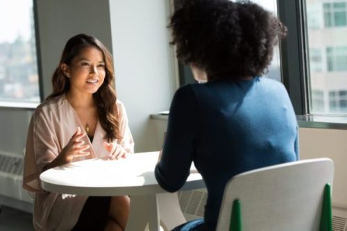 Two women speaking to each other over a table by a window