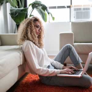 Woman sitting on the floor leaning against a couch working on her laptop