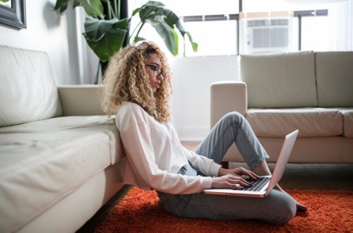 Woman sitting on the floor leaning against a couch working on her laptop
